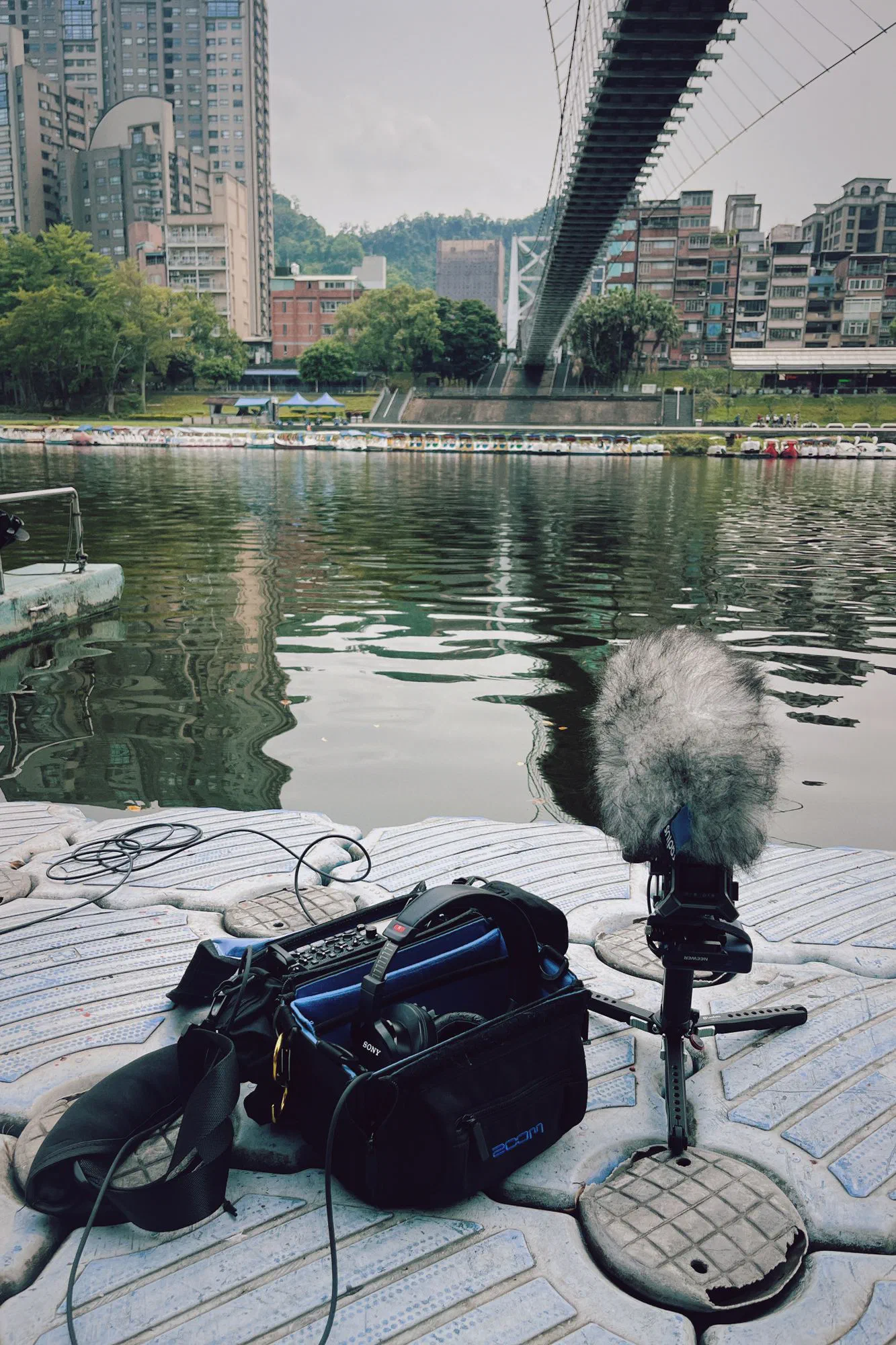 A field recorder in an open bag and a microphone in a fur windshield on a floating dock beside the river, with a suspension bridge and apartment blocks behind.