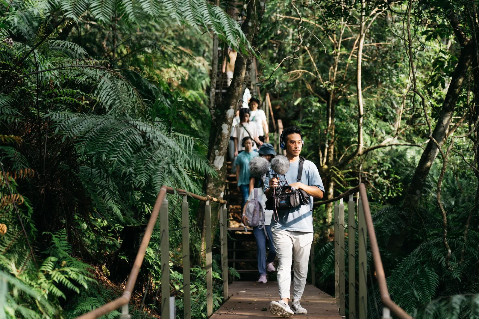 A person carrying a microphone rig and recorder walks along a forest boardwalk, with a line of participants behind.