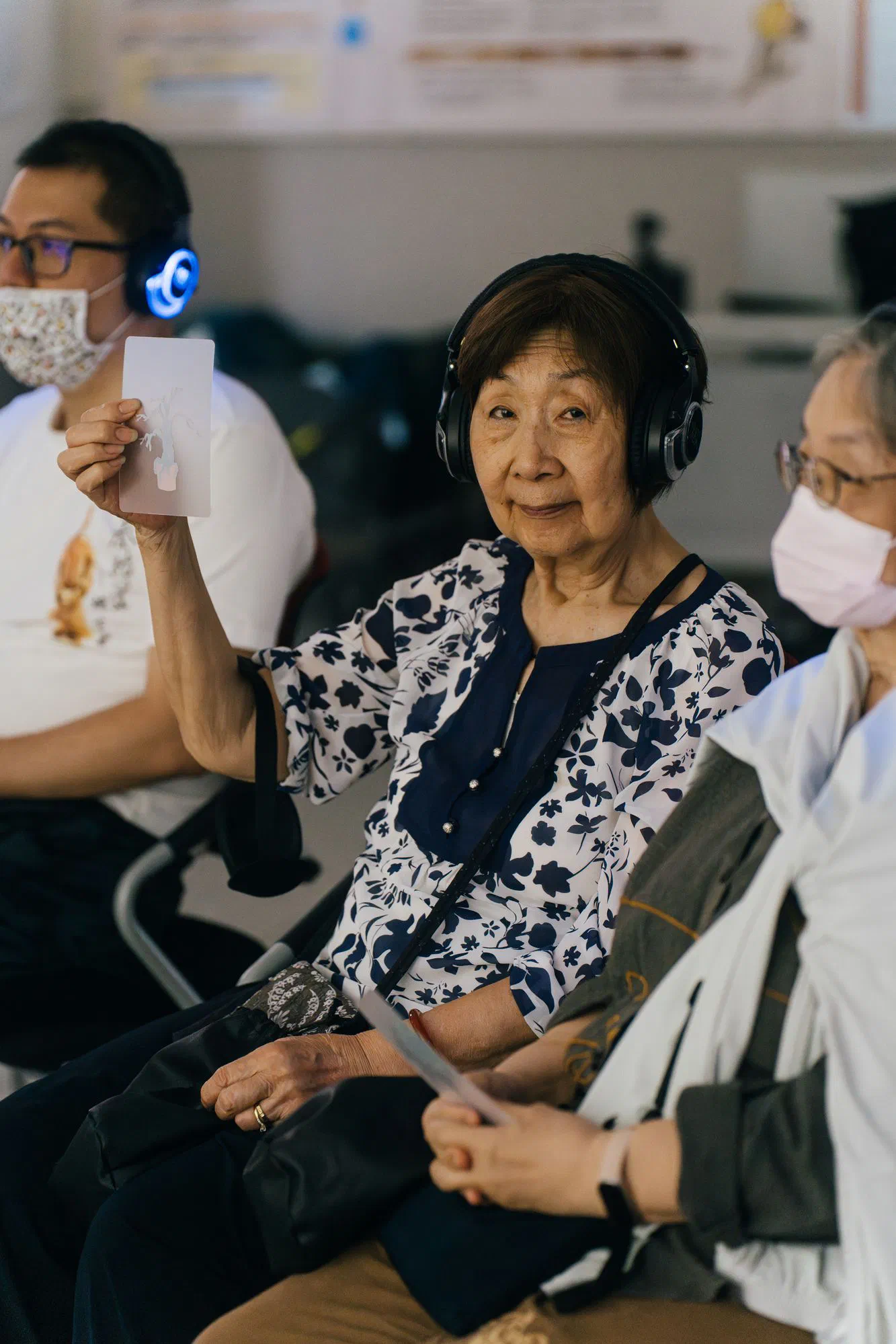 A woman wearing headphones holds up a small printed card and looks toward the camera.