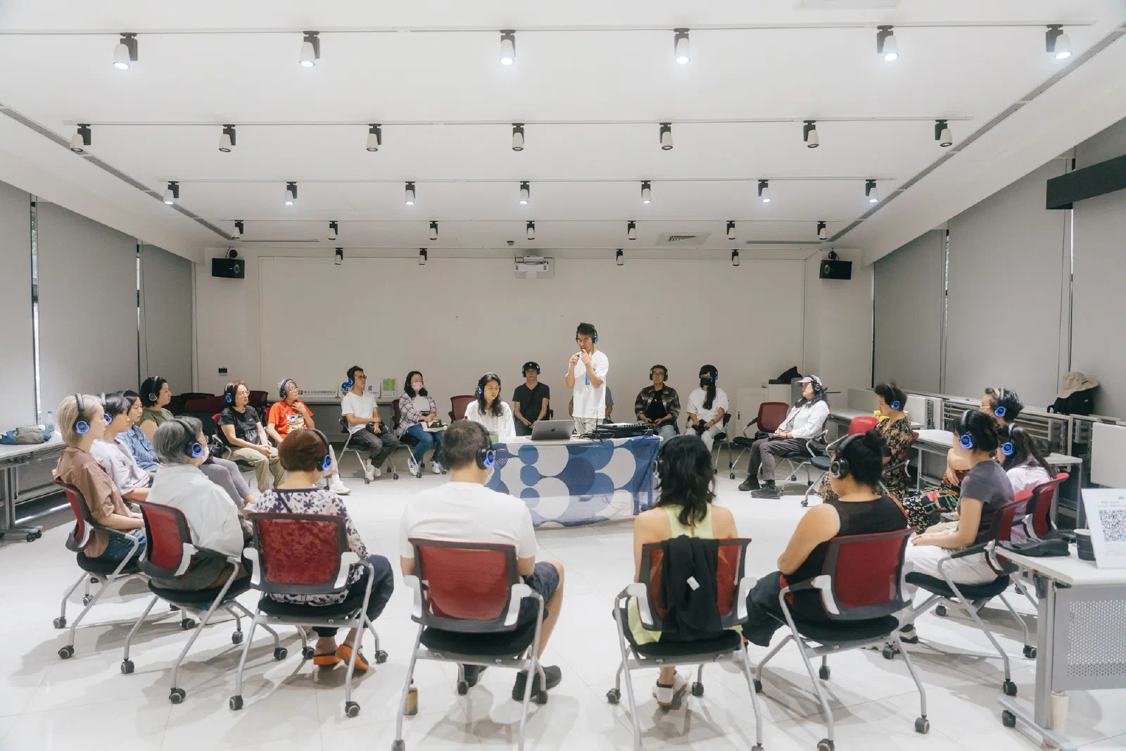 Participants wearing headphones sit in a wide circle of chairs in a bright room, with a person standing at a laptop and mixing table at the centre.