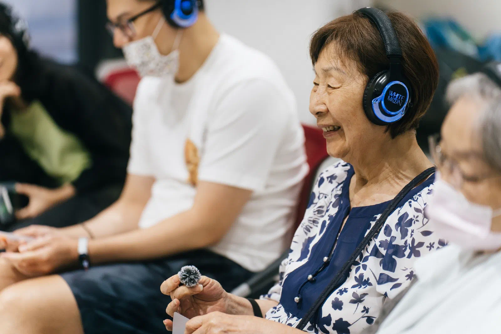A smiling woman wearing illuminated headphones holds a small furry microphone, with other participants seated beside her.