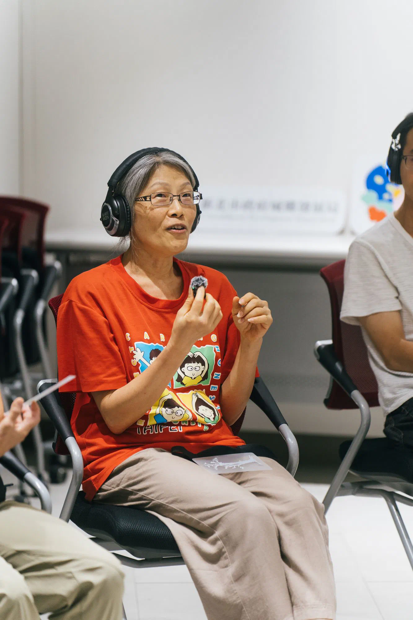 A woman in an orange T-shirt and headphones speaks while holding a small furry microphone, seated in a circle of chairs.