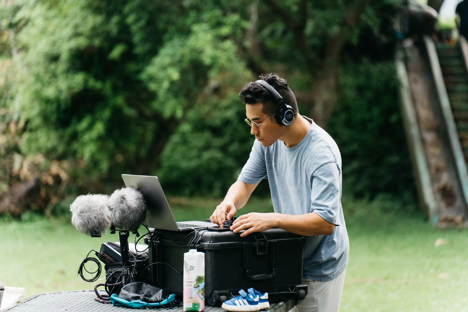 A person in headphones works at a laptop on a black equipment case outdoors, beside two microphones in fur windshields.