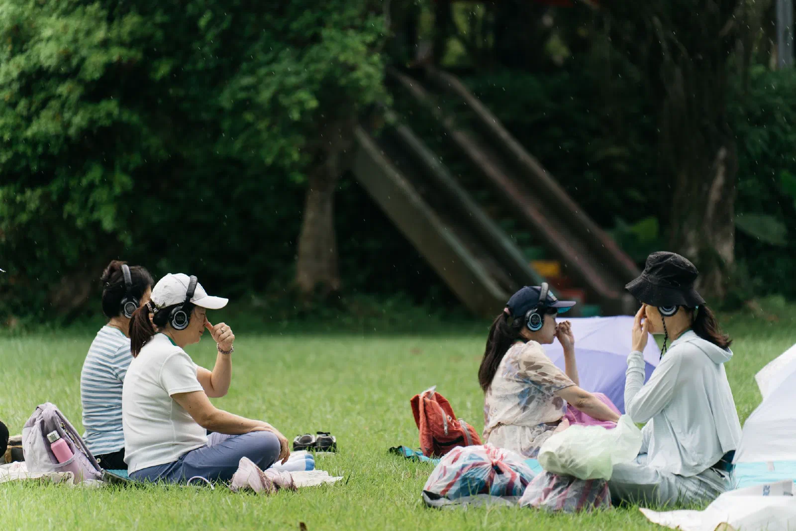 Participants sit on a lawn in headphones, hands raised to their faces, with trees and a slide behind them.