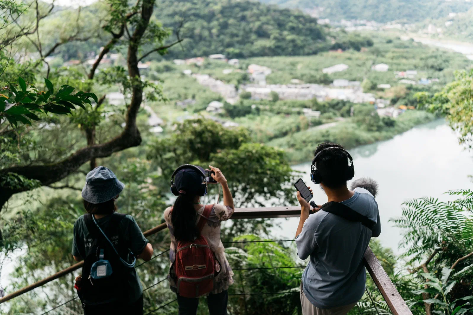 Three participants in headphones stand at a wooden railing, looking down on the river and valley.