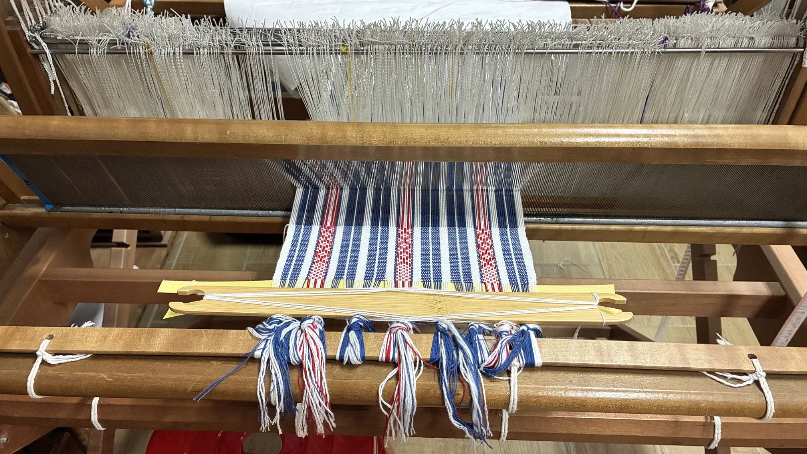 A wooden loom with a half-woven cloth in blue, white and red stripes and bundles of loose thread.