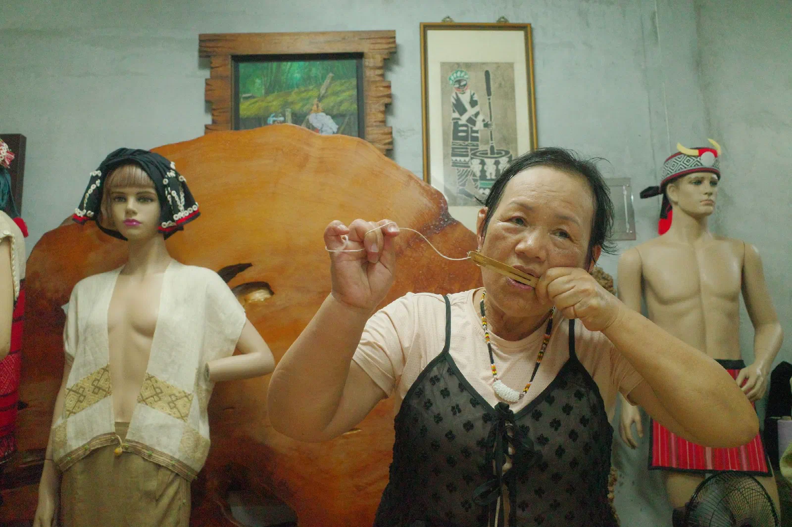 A woman holds a thin cord to a small bamboo instrument at her mouth, with traditional garments on mannequins behind her.