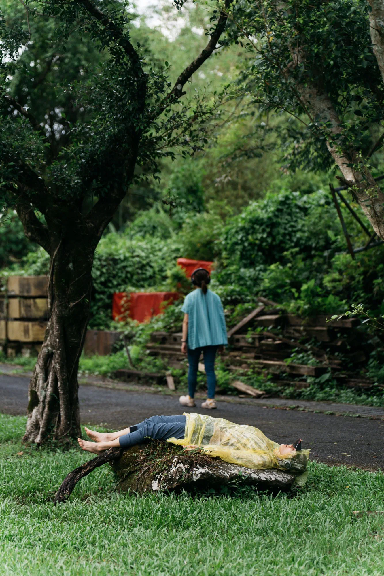 A person in a yellow rain poncho lies on a rock in the grass while another walks away along a path wearing headphones.