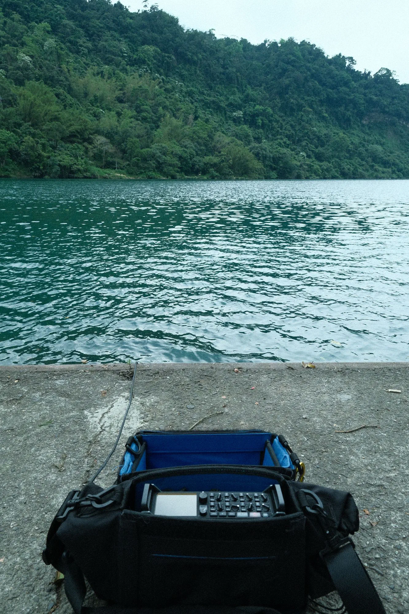 A field recorder in its carrying bag on a concrete embankment, facing a green river and forested hillside.