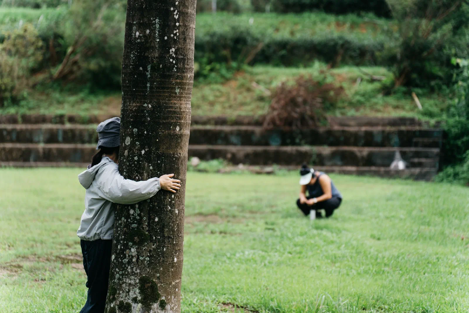 One participant embraces a tree trunk while another crouches on the grass in the distance.