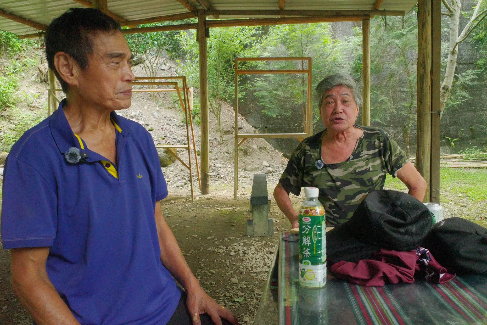 Two elderly men sit in a covered outdoor space during an interview, each wearing a small lavalier microphone.