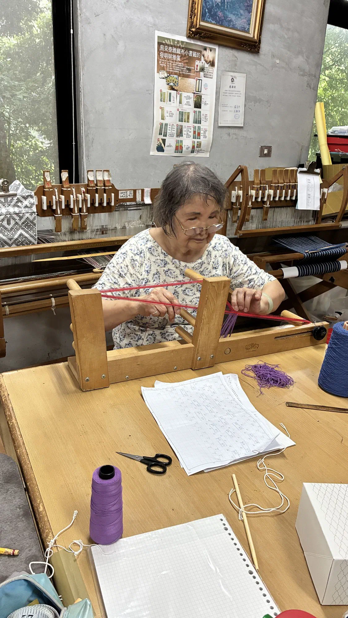 An elderly woman seated at a small wooden frame stretching red and purple thread, with pattern sheets on the table.