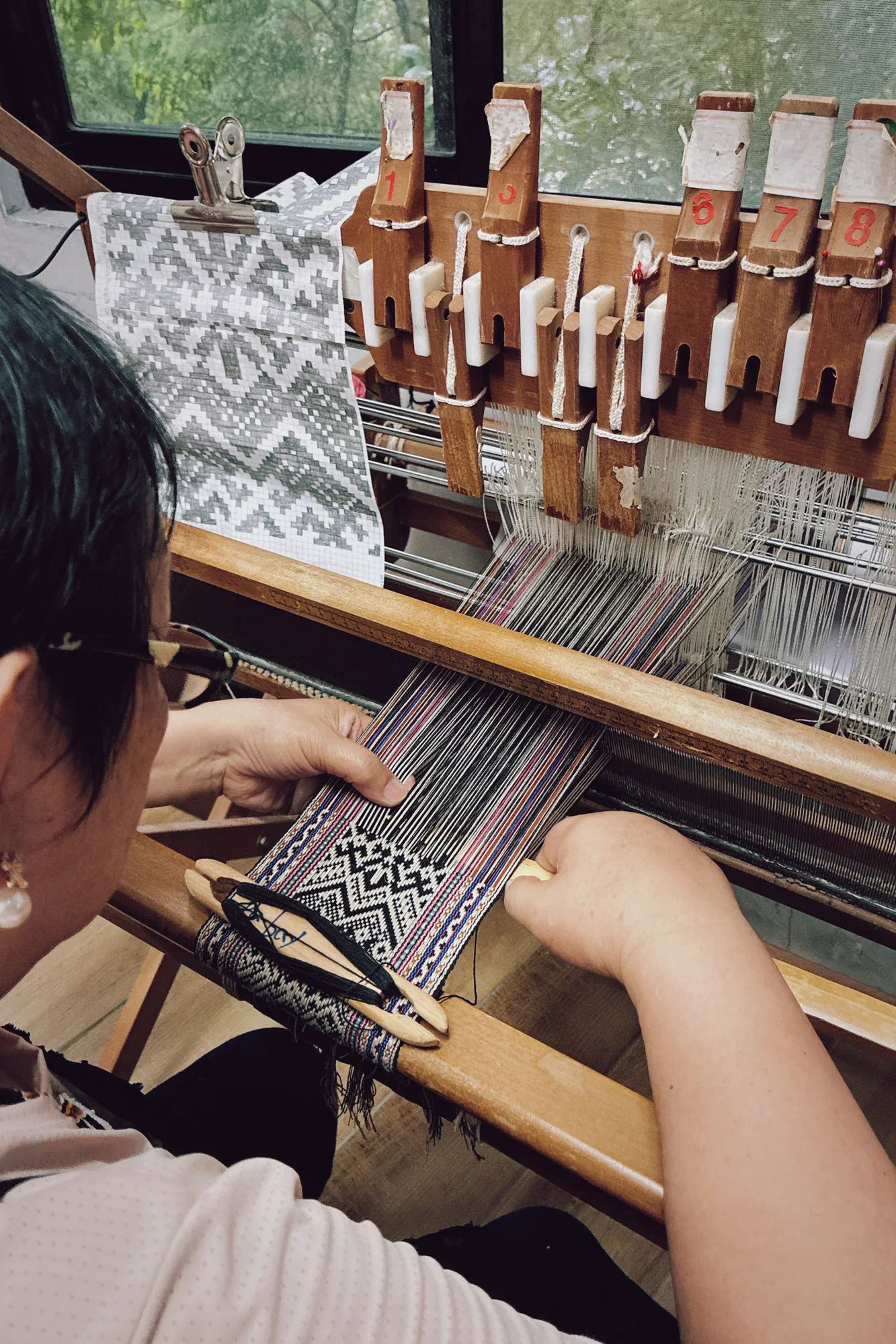 Close view of hands weaving a patterned band on a wooden loom, with a printed pattern chart clipped beside it.