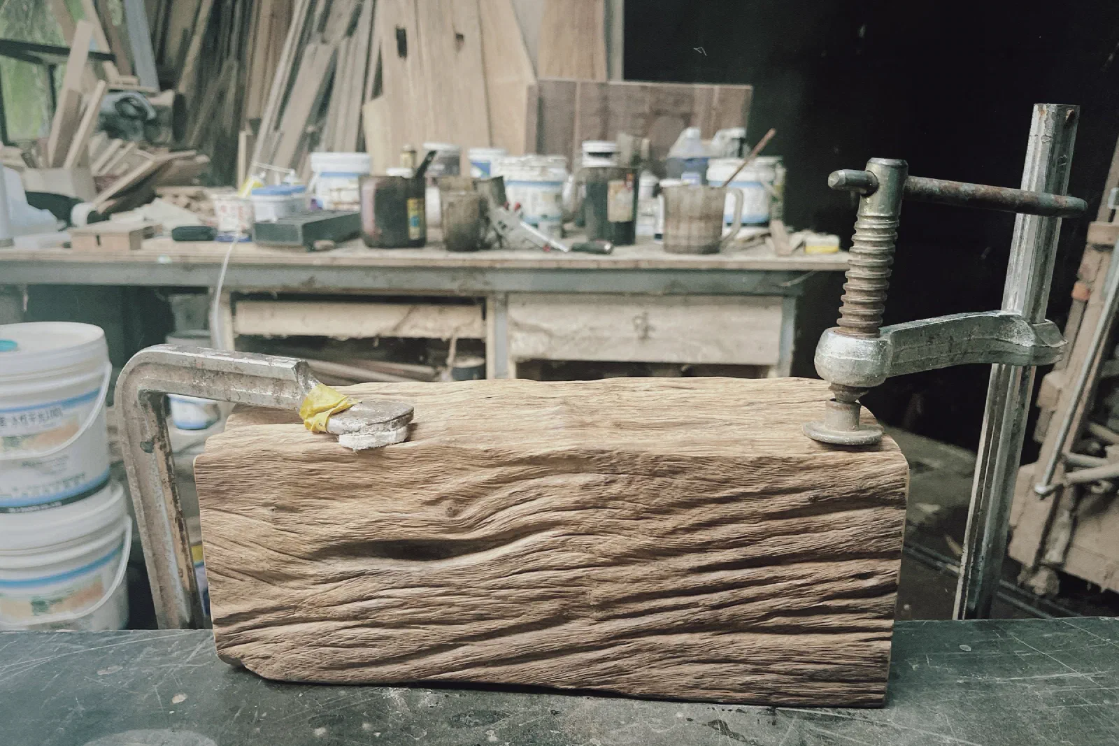 A weathered block of wood with deep grain held by metal clamps on a workbench, with paint tins and timber in the workshop behind.