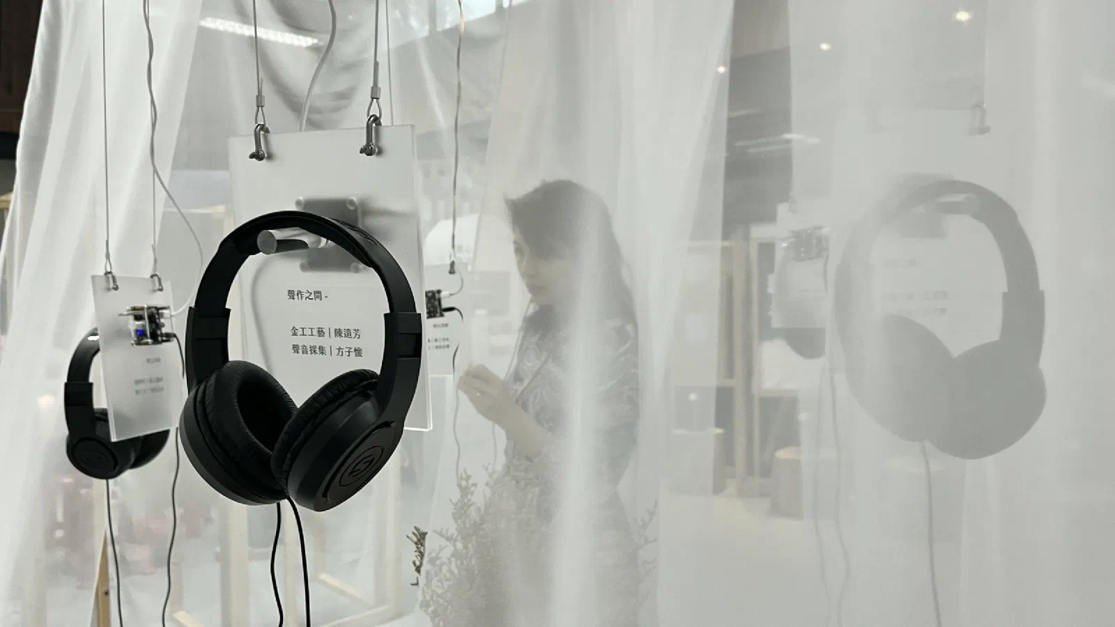 Black headphones hang from wires beside small acrylic labels, seen through translucent white fabric, with a visitor's blurred figure behind.
