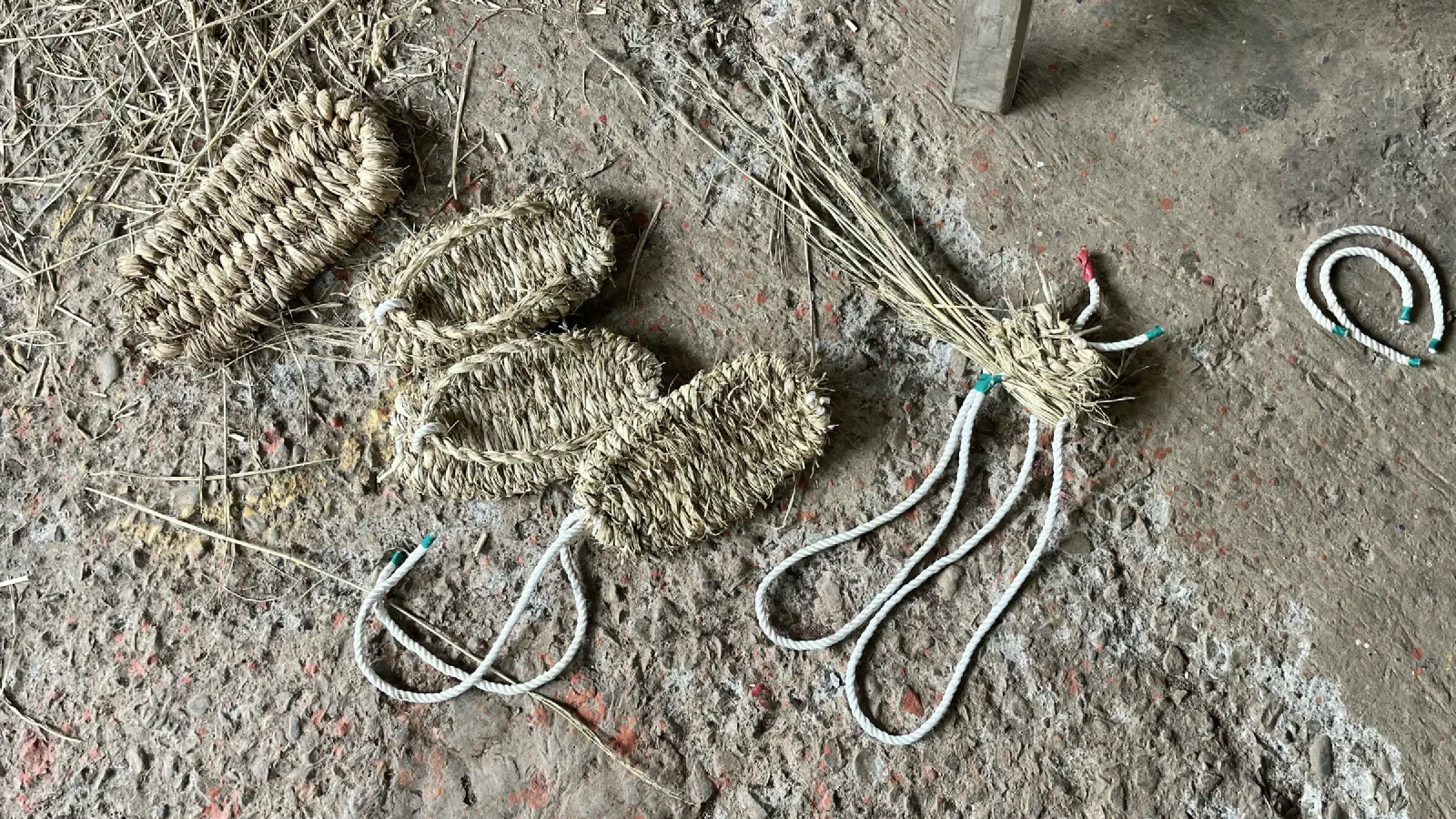Woven grass sandals, a bundle of grass stalks and white cords lie on a stained concrete floor.