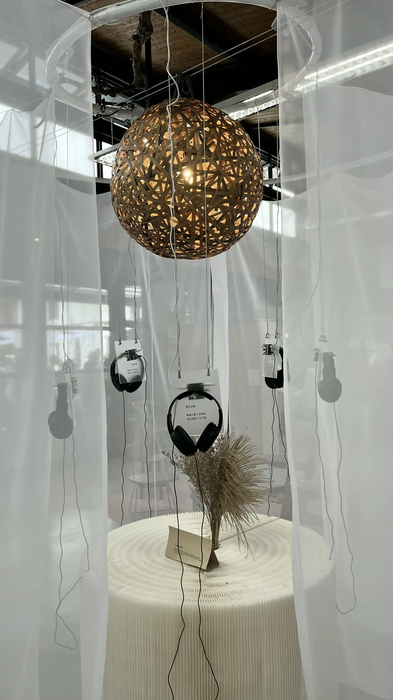 A glowing woven sphere hangs above a round white plinth with dried grasses, surrounded by suspended headphones and sheer white fabric.