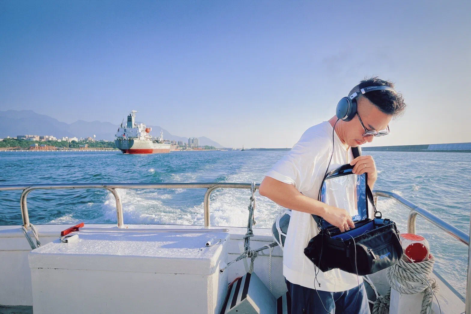 A person in headphones opens a field recorder bag on the back deck of a boat, with a cargo ship and mountains behind.