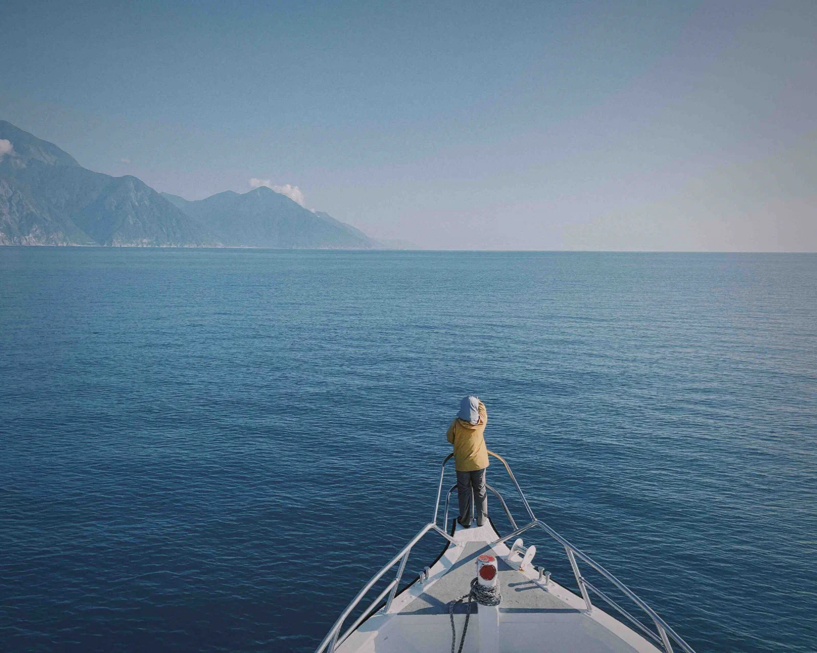 A person in a yellow jacket stands at the bow of a boat facing open water and a mountainous coast.