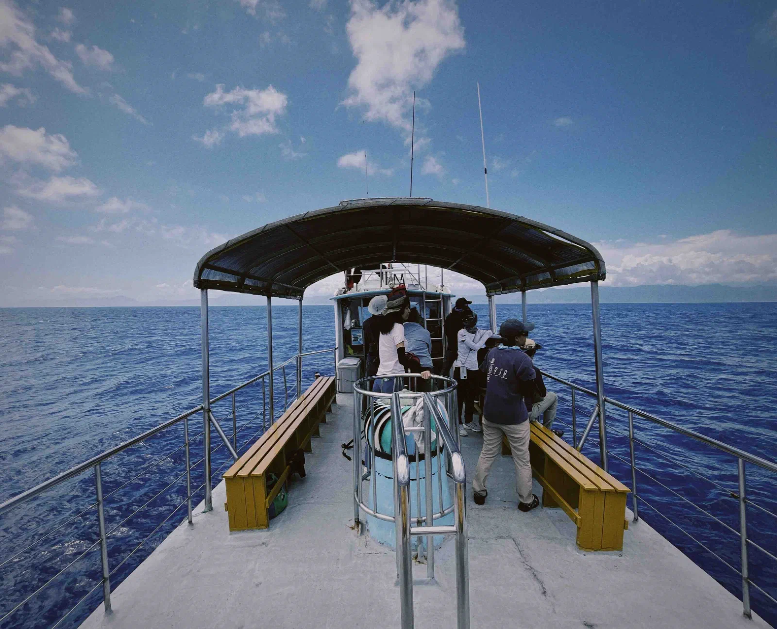 People stand under a canopy on a boat deck, with wooden benches on each side and deep blue sea all around.