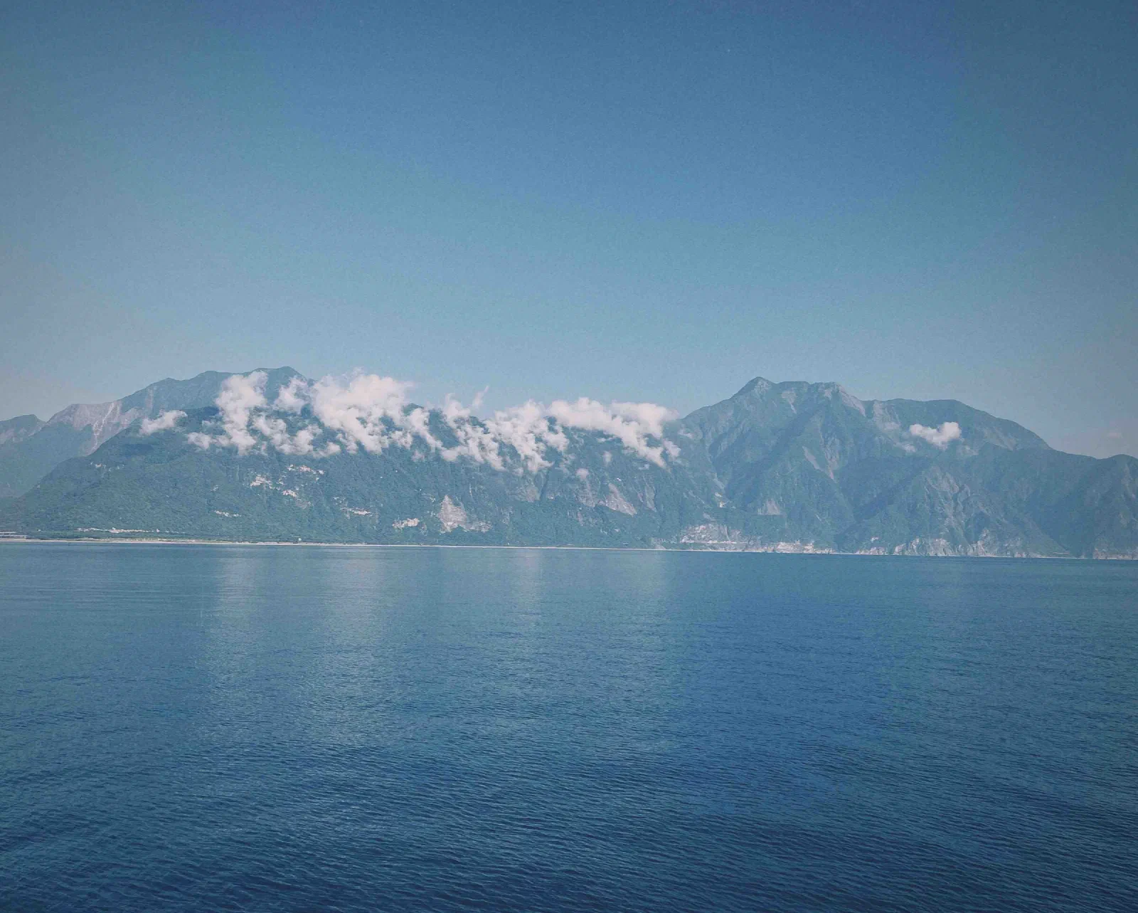 Steep green mountains with low clouds rise behind a wide, calm stretch of blue sea.