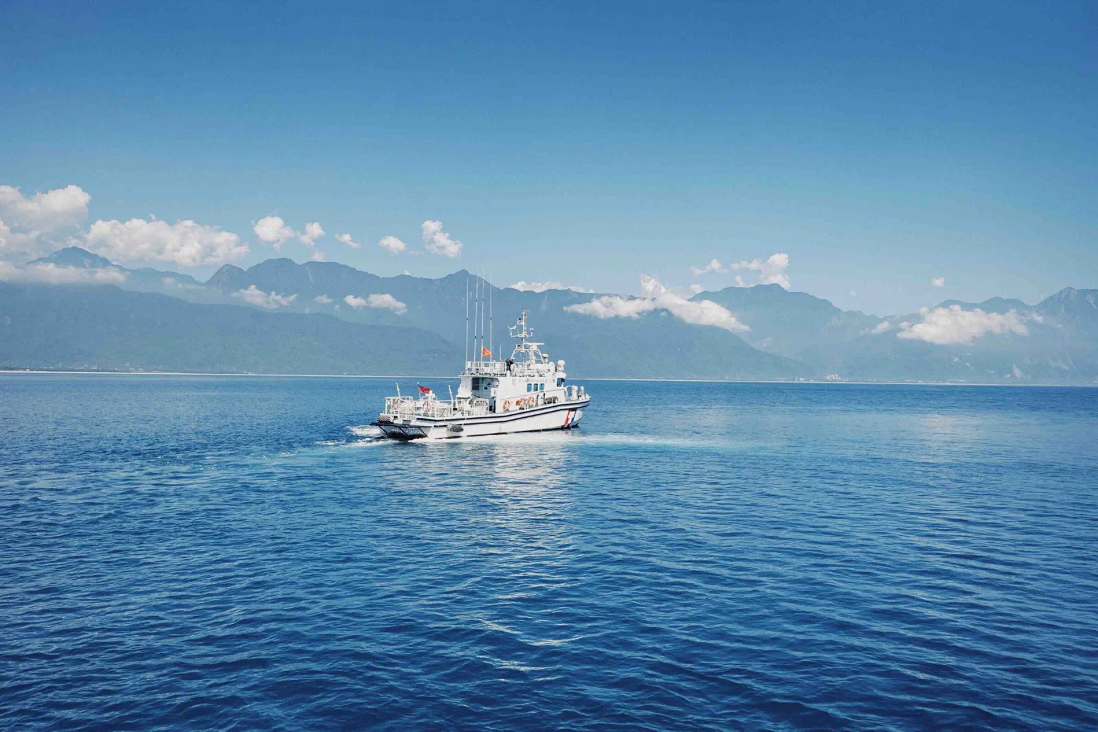 A white patrol boat travels across calm blue sea with a mountain range and clouds behind it.