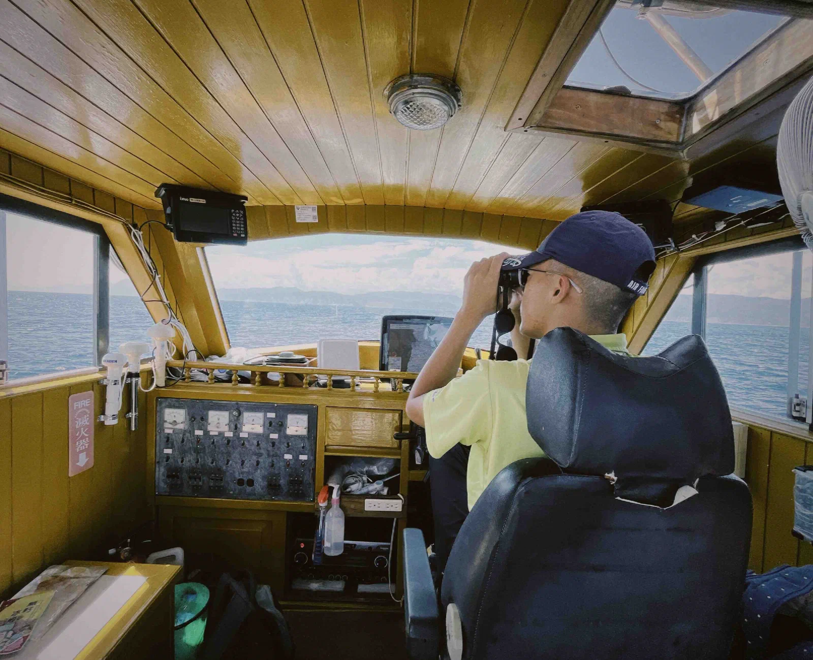 A person in a cap looks through binoculars from a boat's wooden wheelhouse toward the sea.