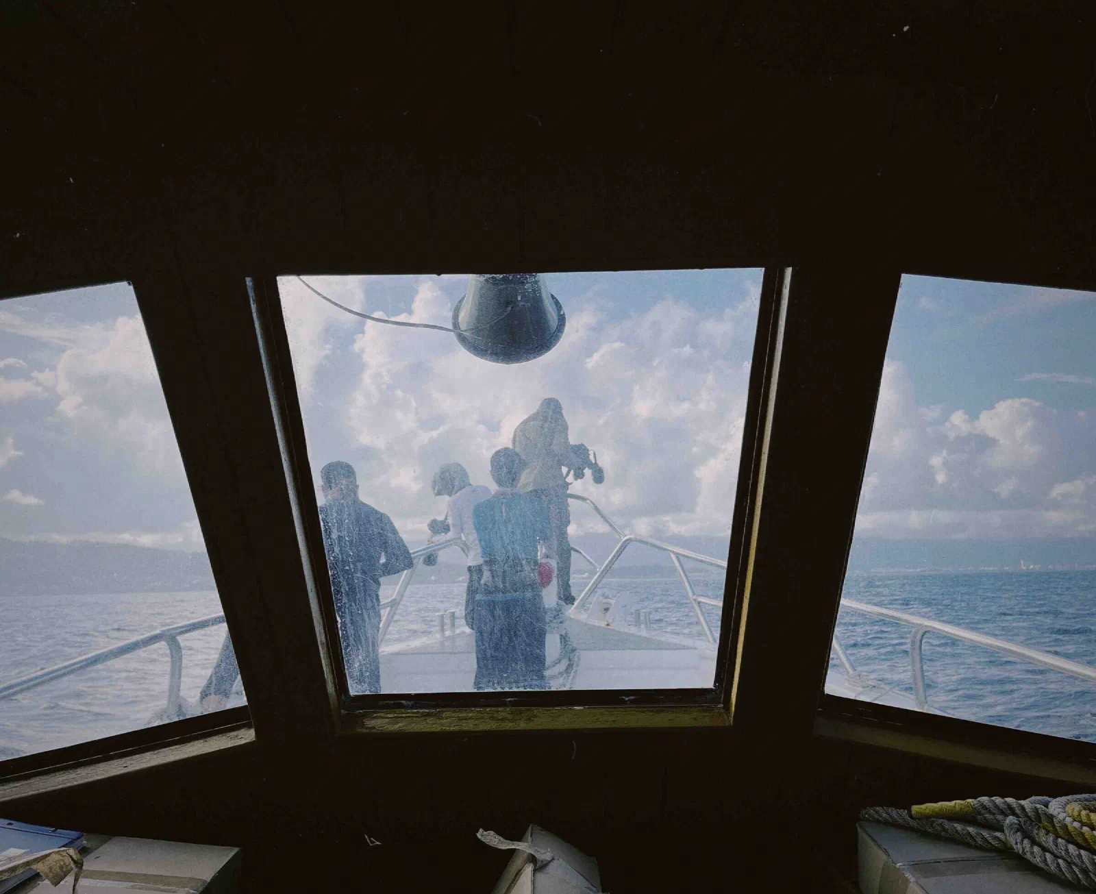 Through a scratched cabin window, several people stand on the bow of a boat facing open sea, one holding a recorder.