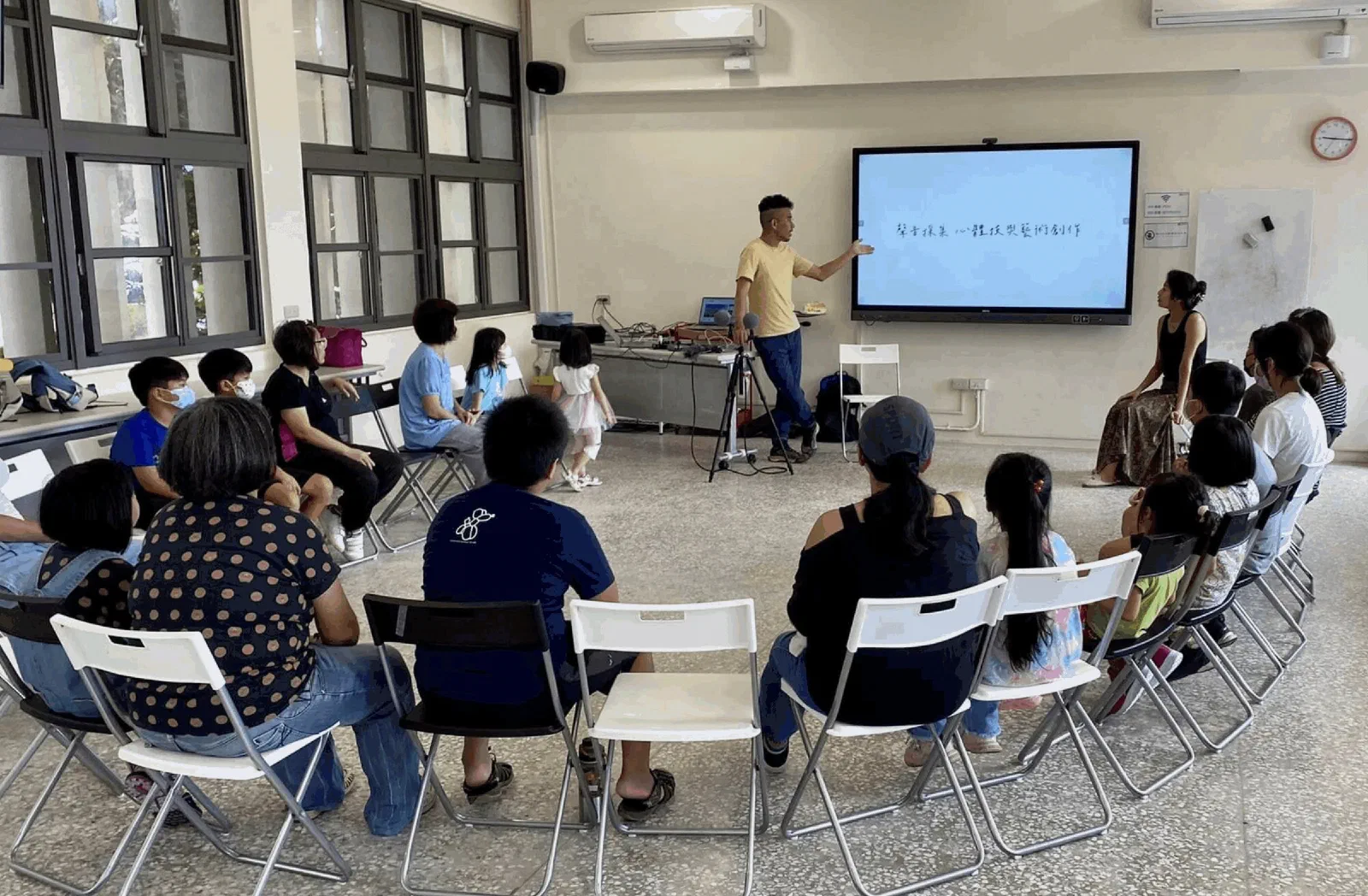 Children and adults sit in a wide circle of folding chairs in a bright classroom while a facilitator in a yellow shirt gestures to a screen, with microphones on a stand and a laptop on a table behind him.