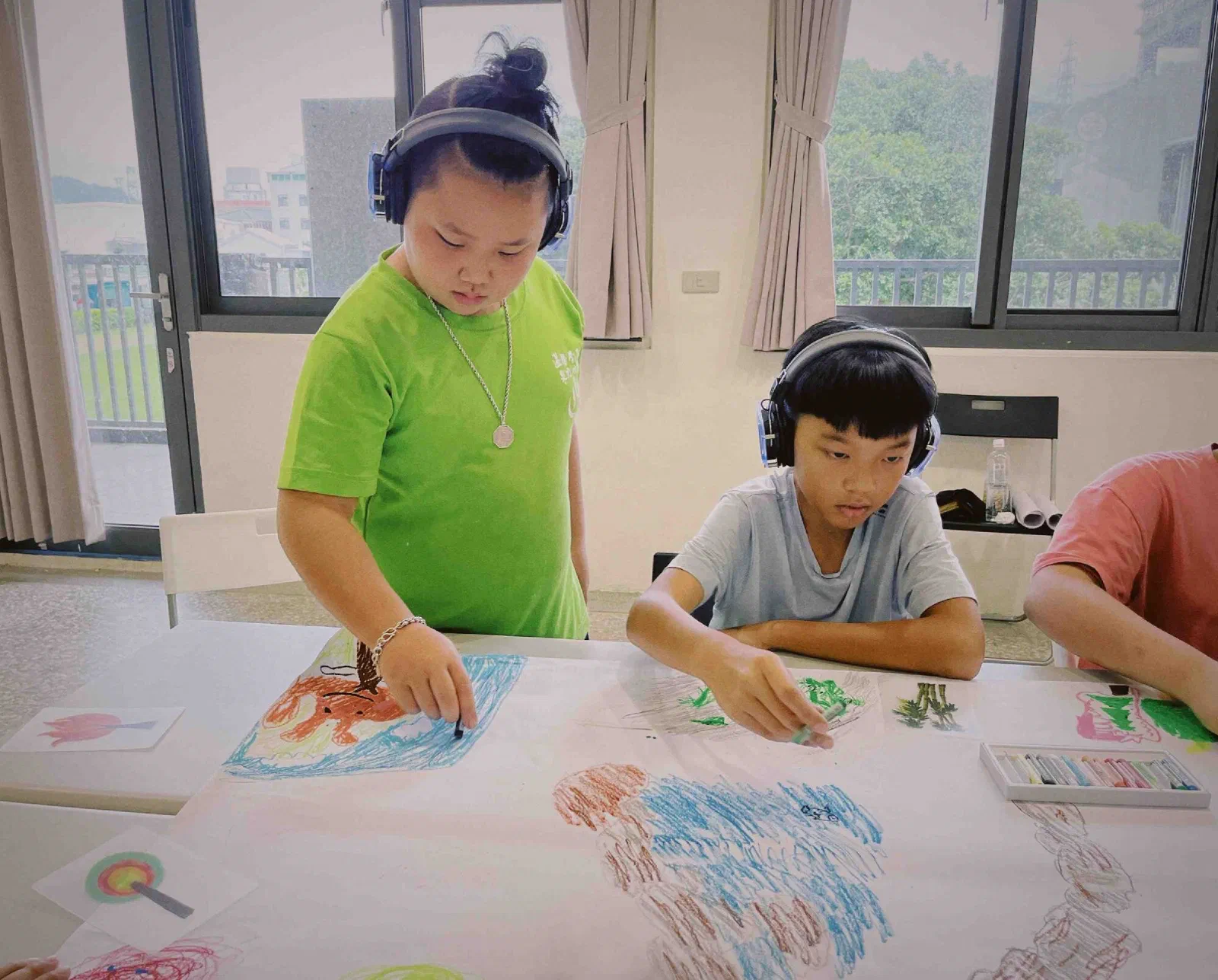 Two children in headphones lean over a long sheet of paper on a table, drawing with oil pastels beside a window.