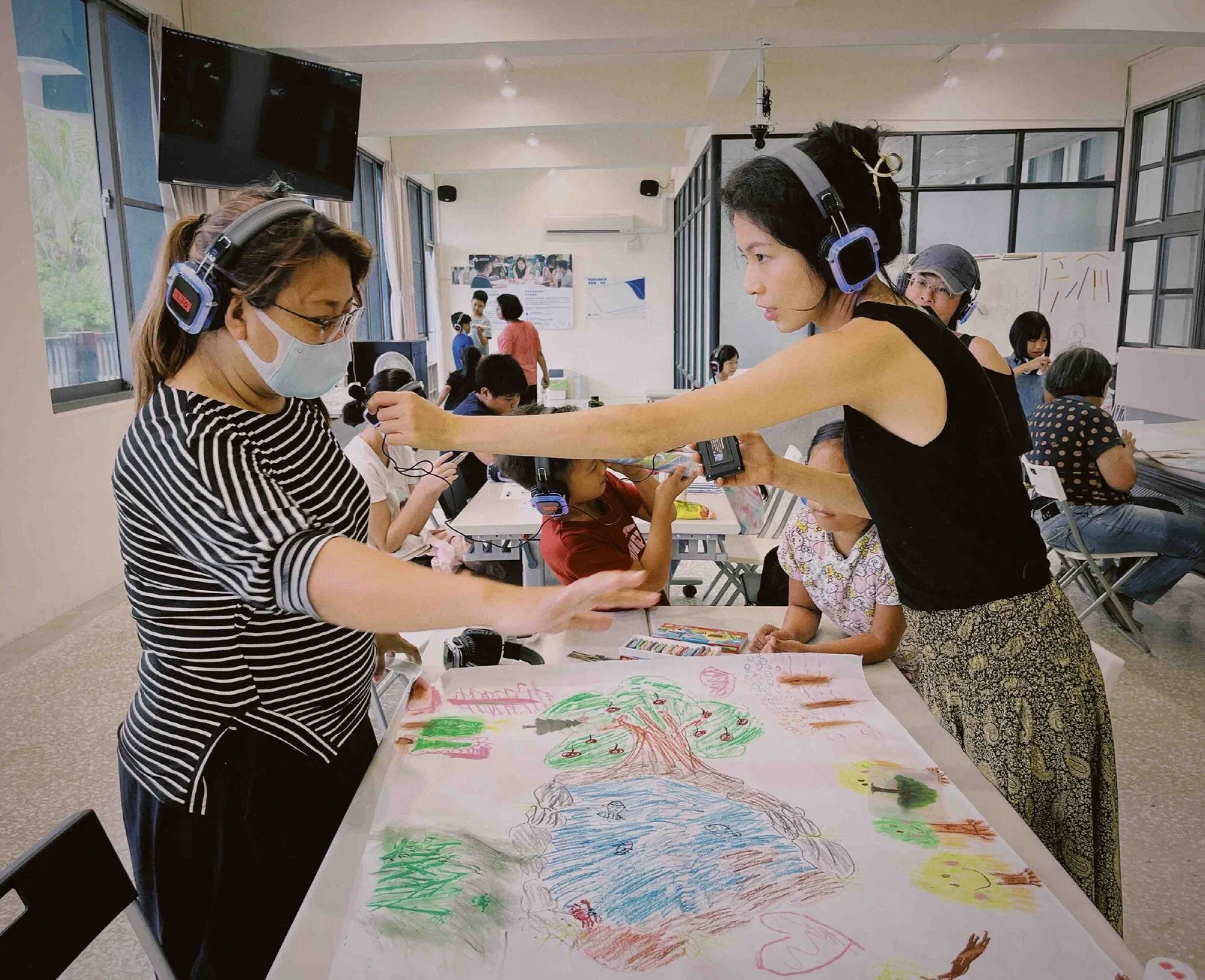 A facilitator in headphones leans across a table to adjust a clip microphone on an adult, with children drawing on a large pastel mural in a bright room behind.