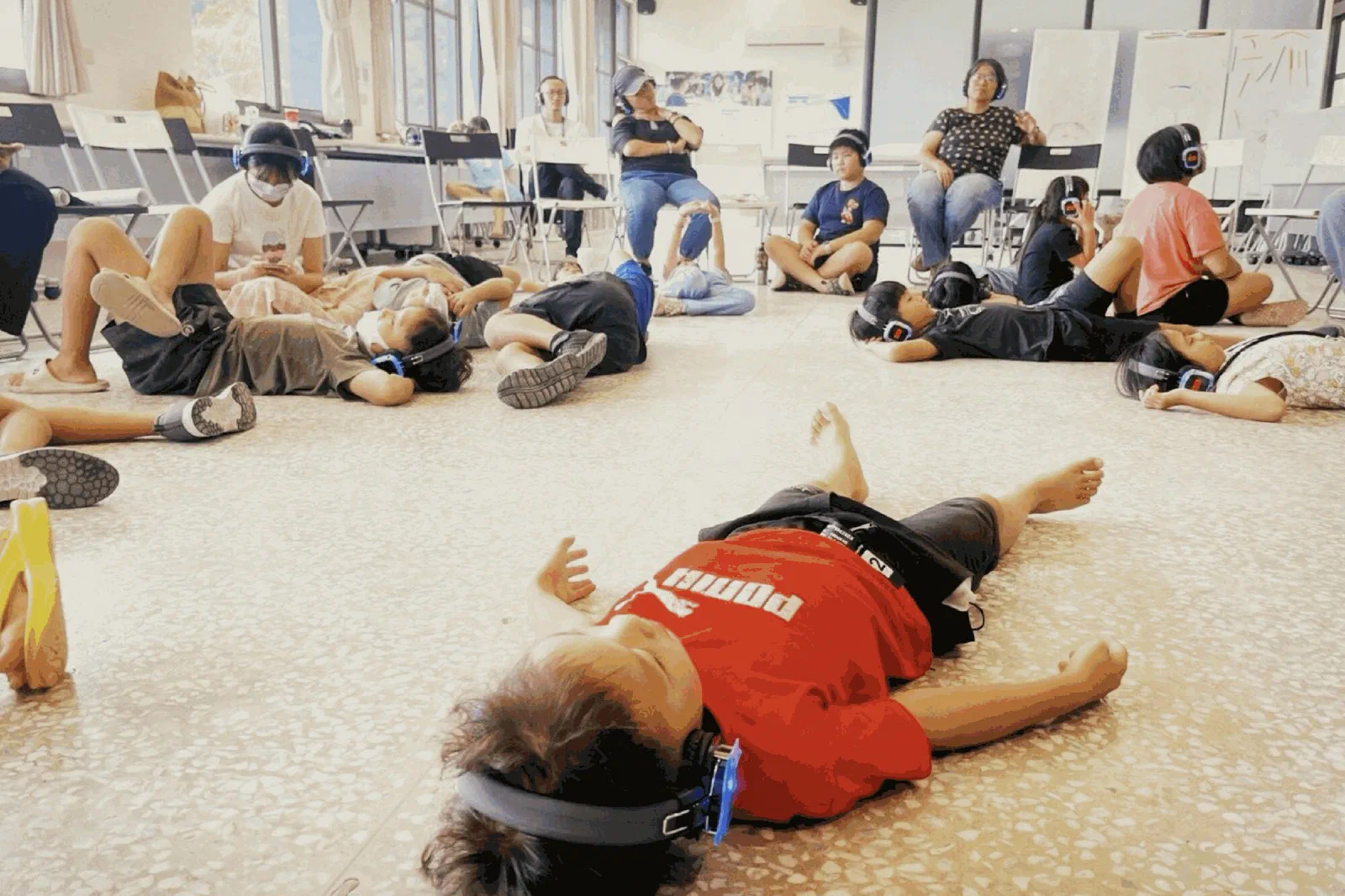 Children lie on the floor wearing headphones, one small child in a red shirt in the foreground with arms outstretched, while adults sit on chairs in the background.