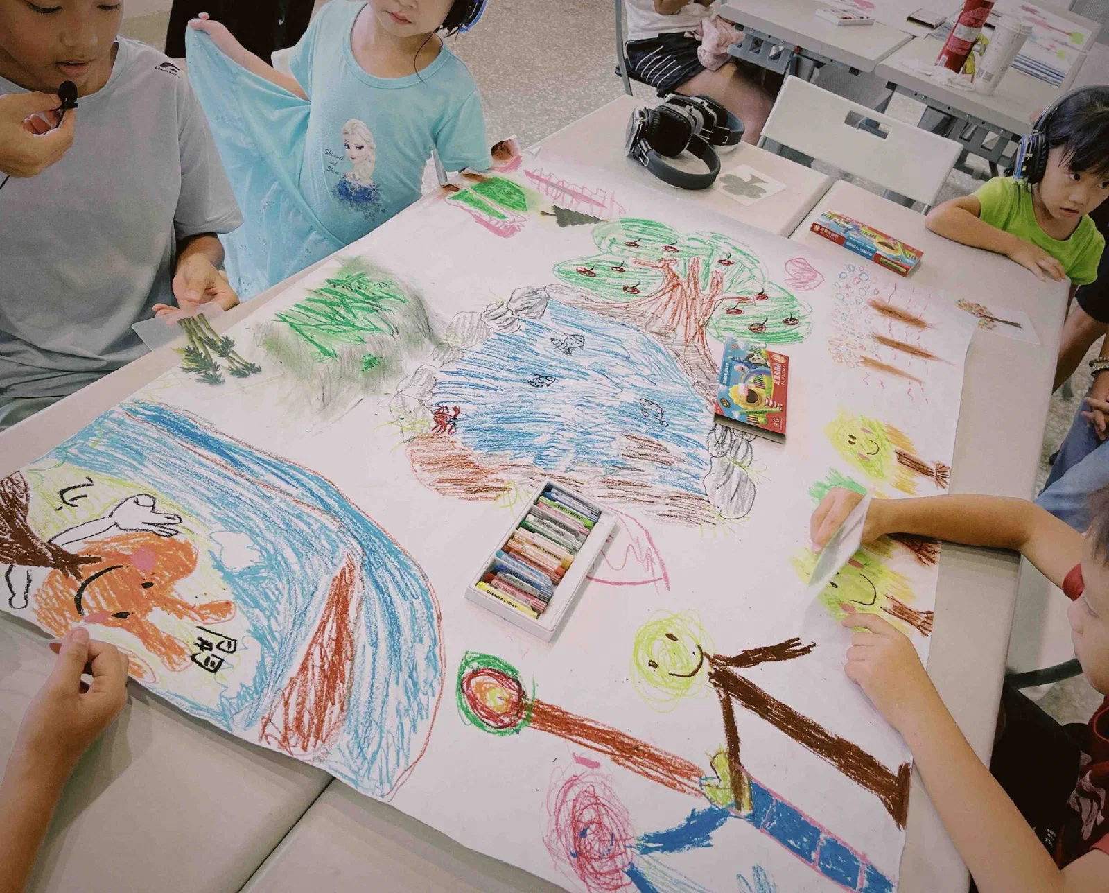 Looking down at a long sheet of paper covered in children's oil-pastel drawings of a waterfall, a pool, trees, a bear and smiling suns, with children around the table.