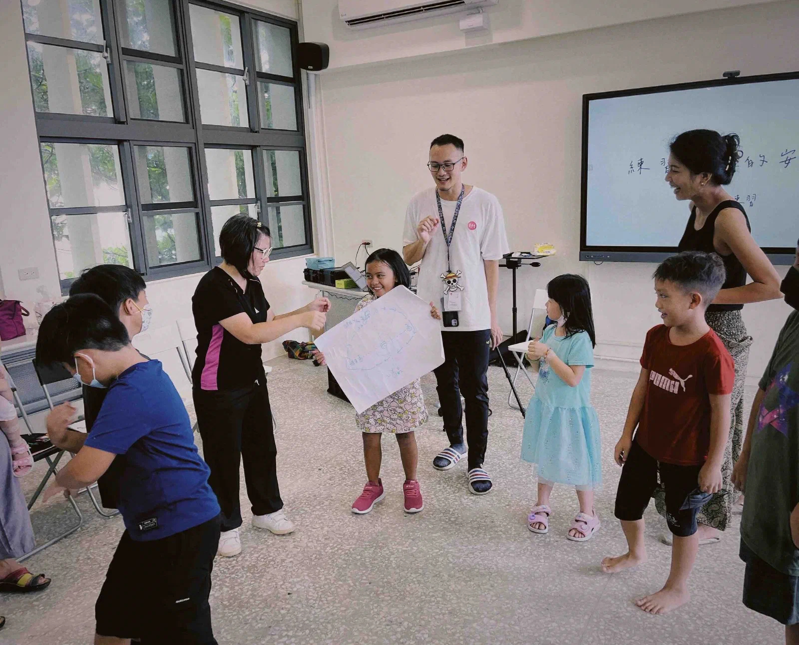 Children stand and move in a bright classroom while an adult holds up a large sheet of paper with a hand-drawn diagram, with a screen behind them.