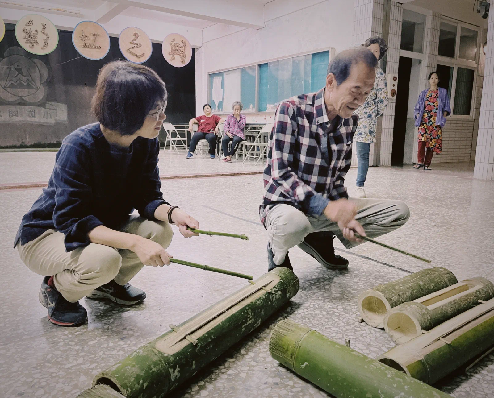 Two people crouch and strike split bamboo tubes with thin sticks in a school hall, while others sit and watch behind them.