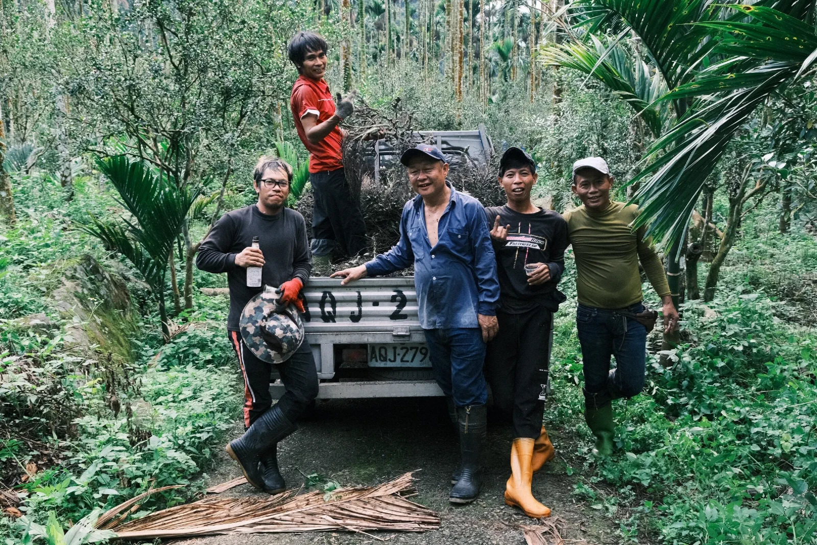 Five workers pose beside a small truck loaded with areca fronds in a green grove, in rubber boots, one holding a bucket hat.