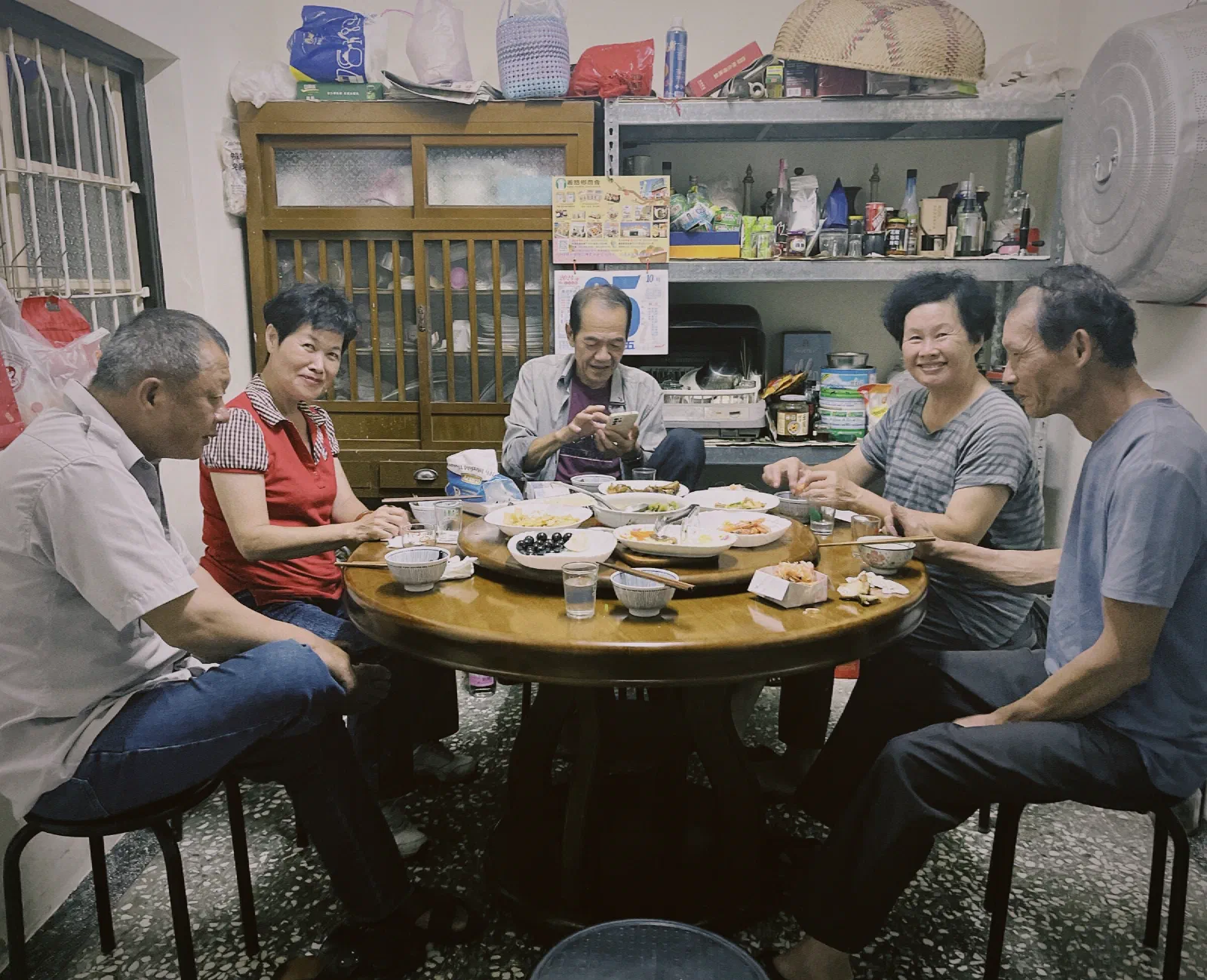 Five villagers sit around a round table with dishes at home, two of them smiling at the camera, with a wooden cabinet and shelves of jars behind.