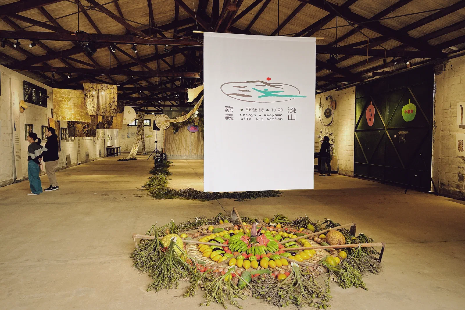 Inside a former ammunition depot with timber roof trusses, a large white banner for the Chiayi Asayama Wild Art Action hangs above a circular arrangement of fruit, vegetables and plants on the floor, with visitors and hanging textile works along the walls.