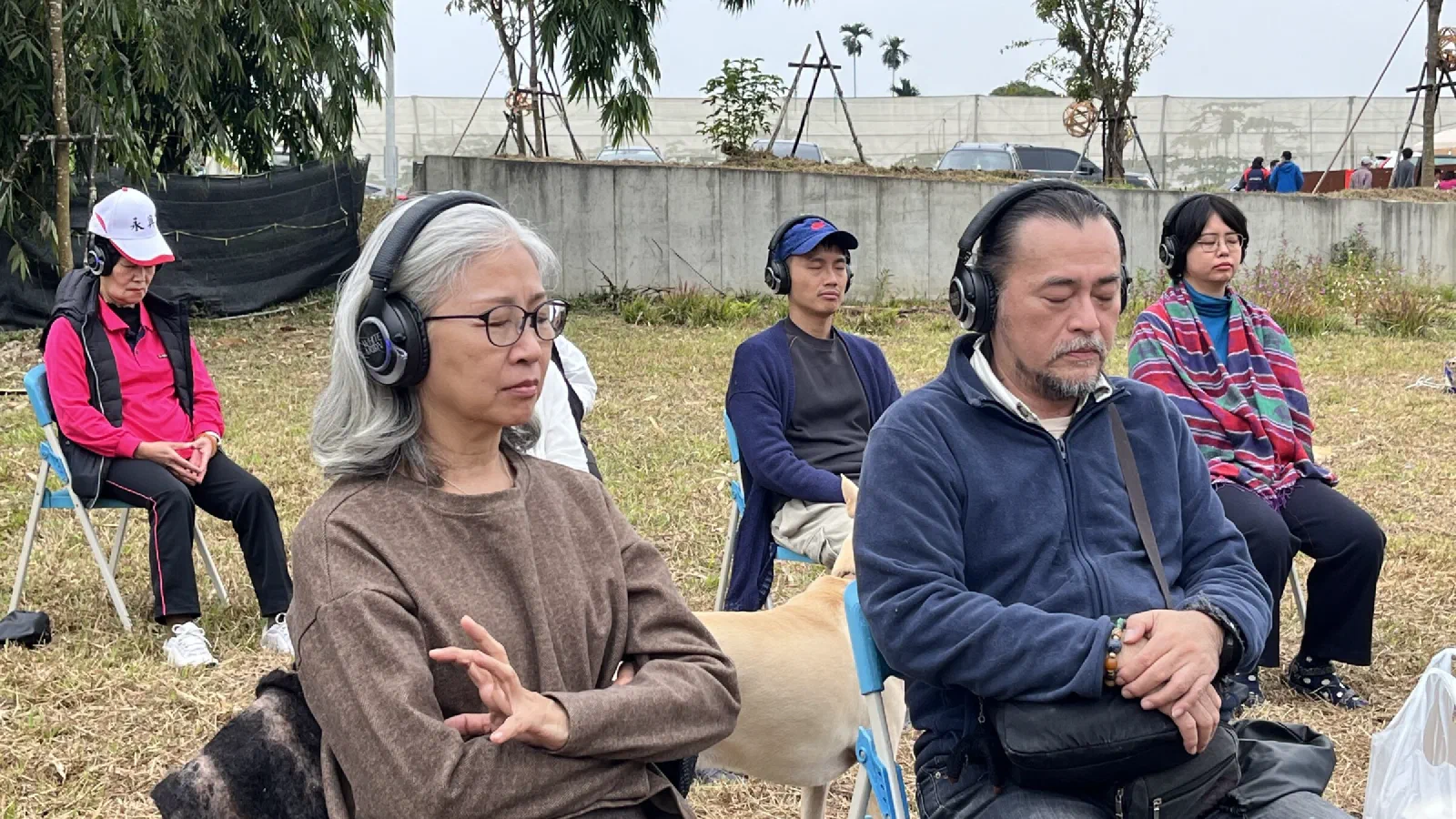 Four participants sit with eyes closed on a grassy field wearing headphones, with a light-coloured dog beside them and a concrete wall behind.