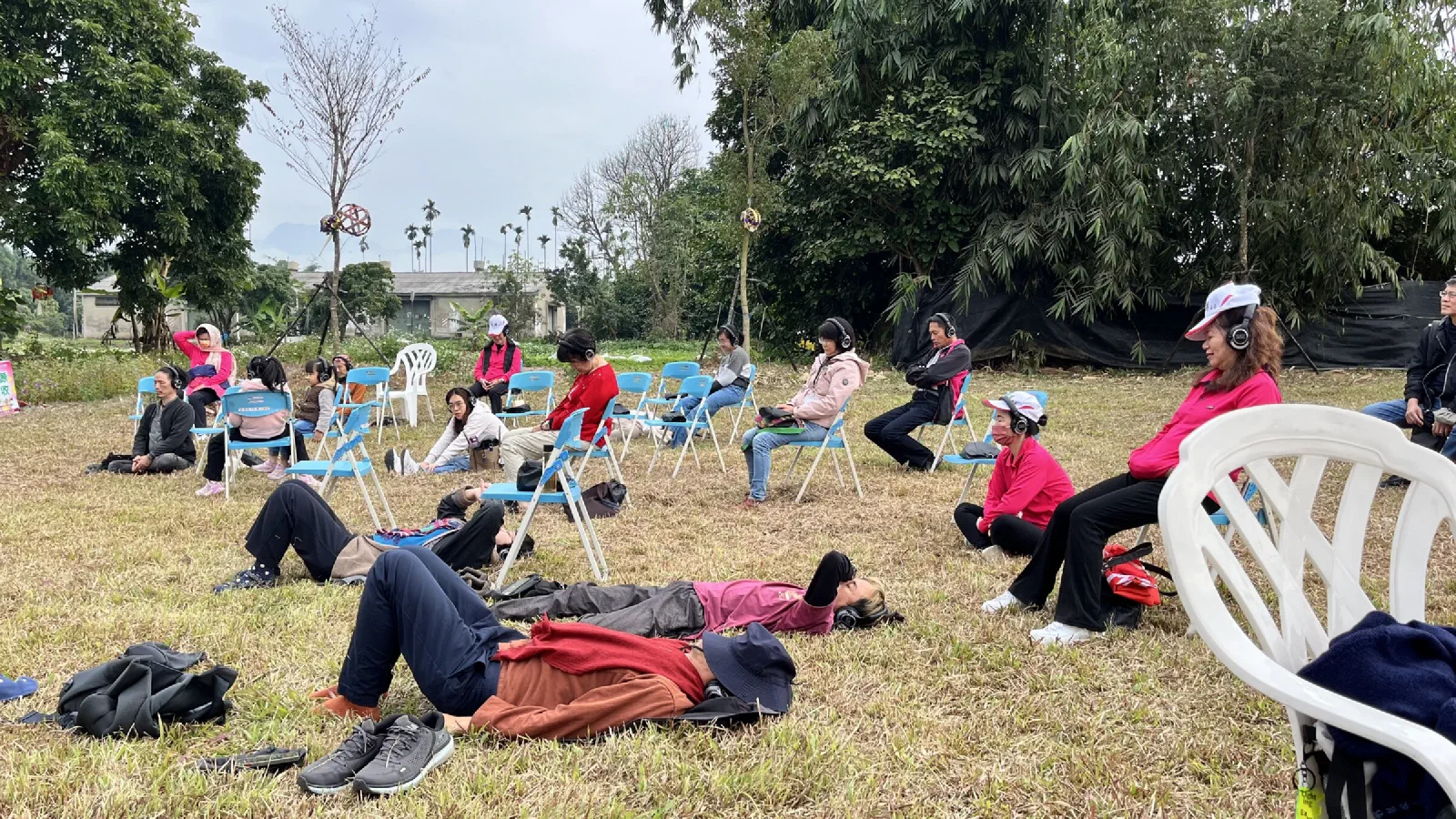 Participants in headphones sit on folding chairs and lie on the grass of an open field, listening with eyes closed, with trees and slender kinetic sculptures behind.