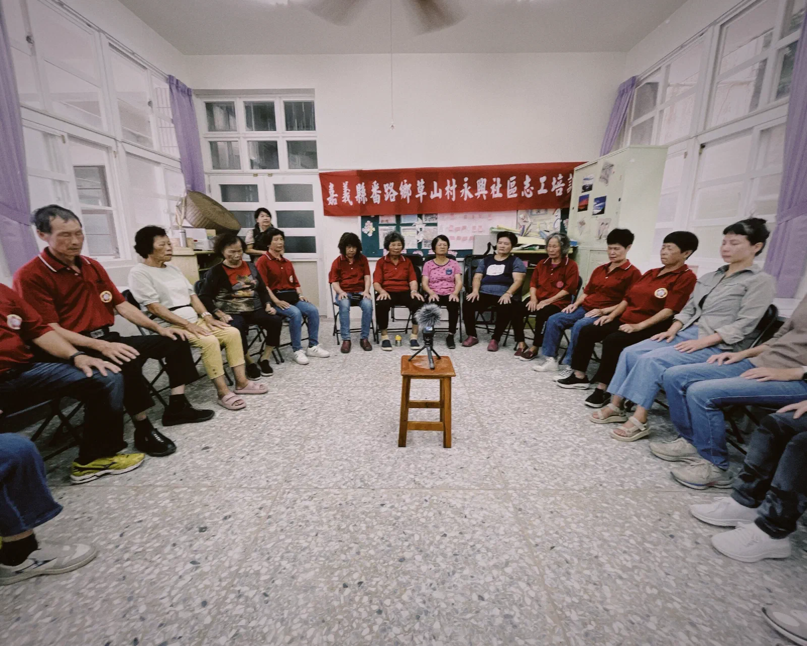 Residents in red shirts and other clothes sit in a wide circle in a community room with eyes closed and hands on their knees, a microphone on a stool at the centre and a red banner behind.