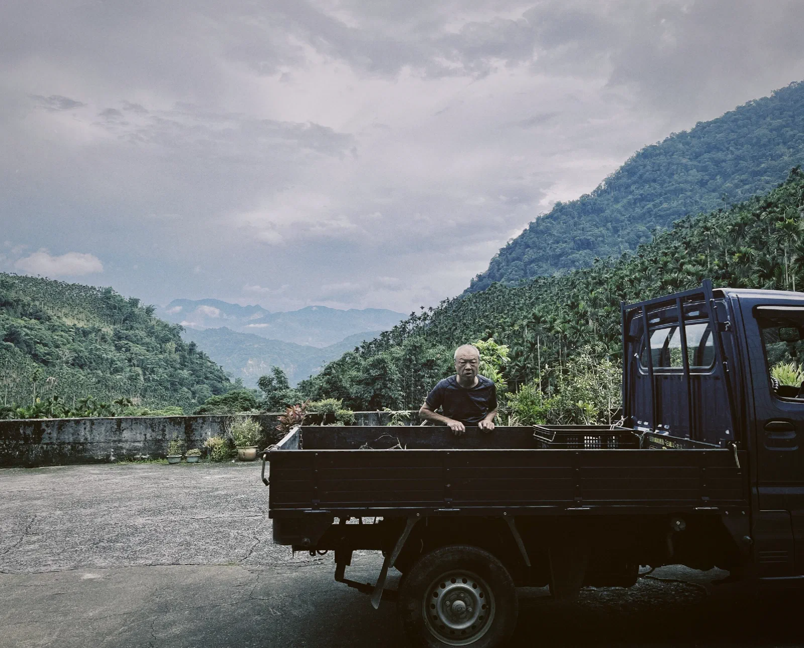 A man looks over the side of a dark pickup truck on a concrete yard, with areca-covered mountains and cloudy sky behind.