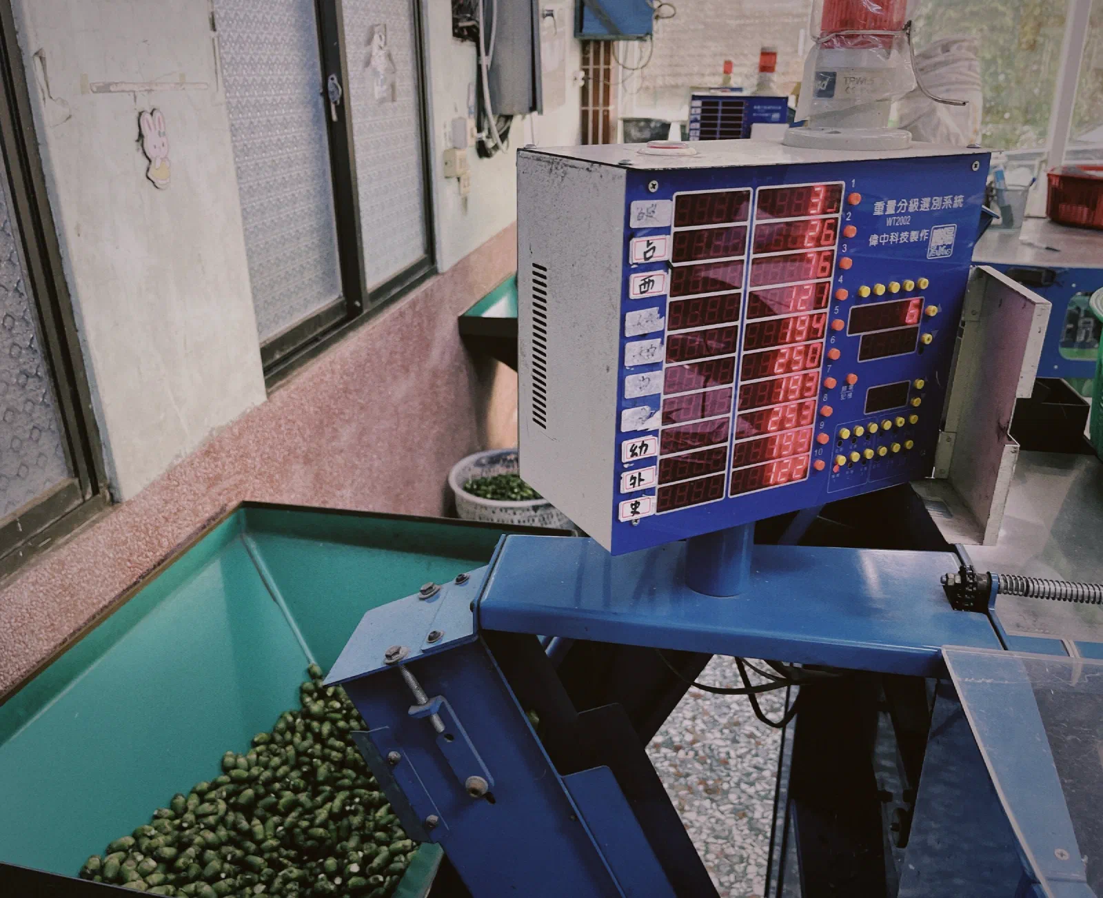 A round blue sorting machine with numbered chutes and hoses leading into large sacks of areca nuts, with handwritten paper slips on top and small microphones attached to its side.