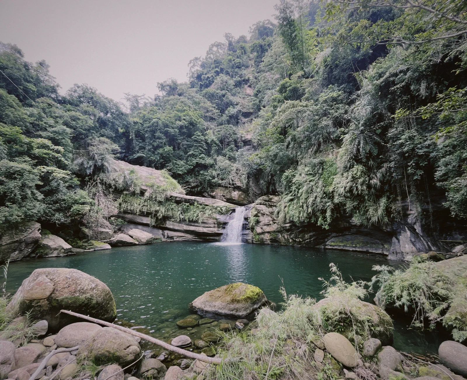 A small waterfall drops into a deep green pool among mossy boulders and steep, fern-covered rock walls.