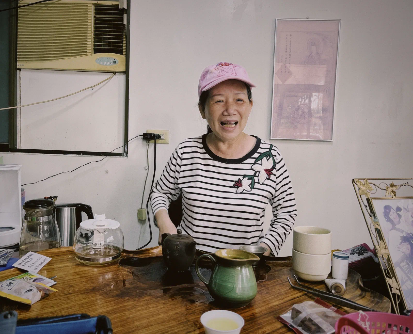 A smiling woman in a pink cap and striped top sits behind a wooden tea table with a teapot, pitcher and cups, an air conditioner and framed picture on the wall behind her.