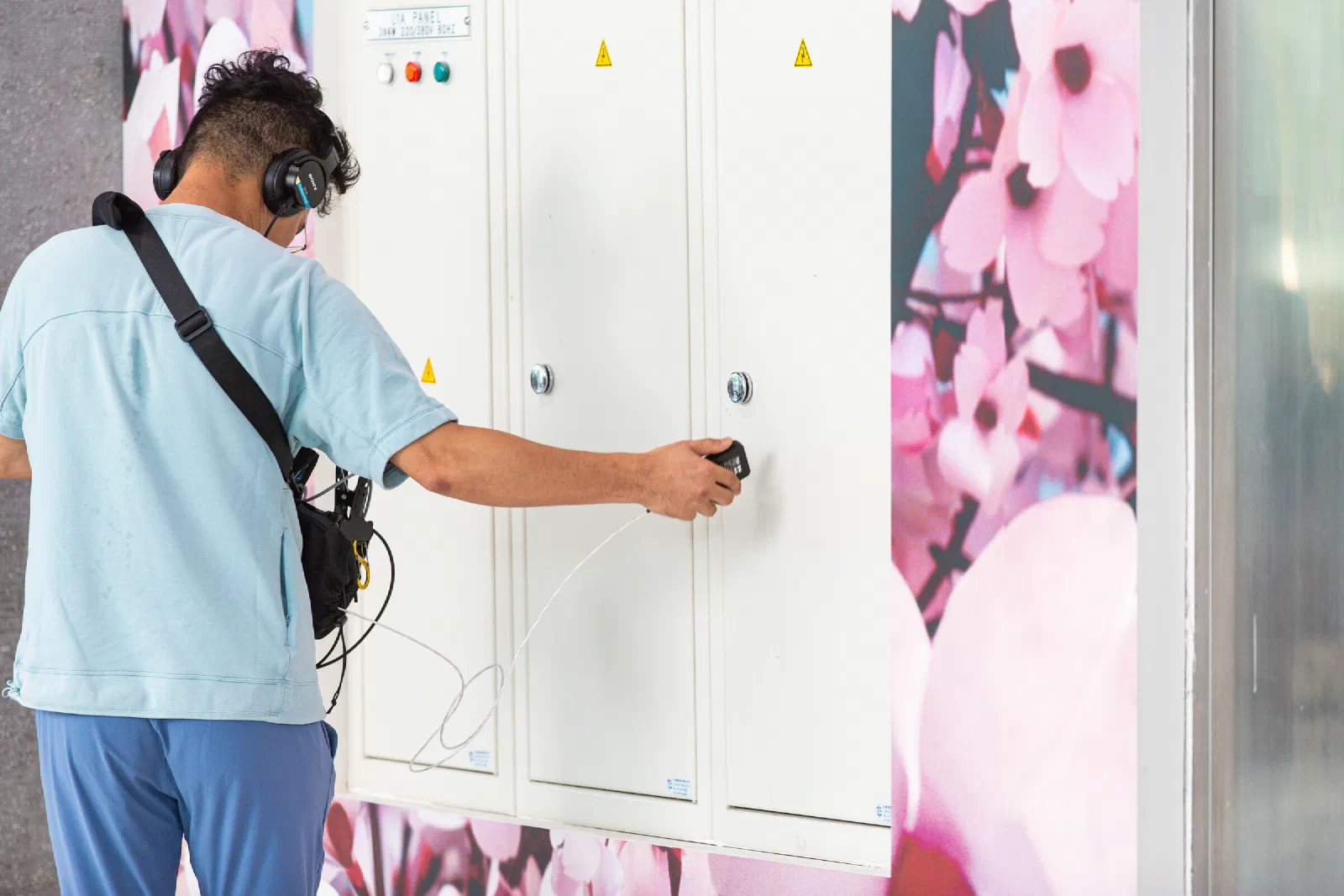 A person in headphones, seen from behind, holds a small contact microphone against a white electrical cabinet in front of a pink floral wall.