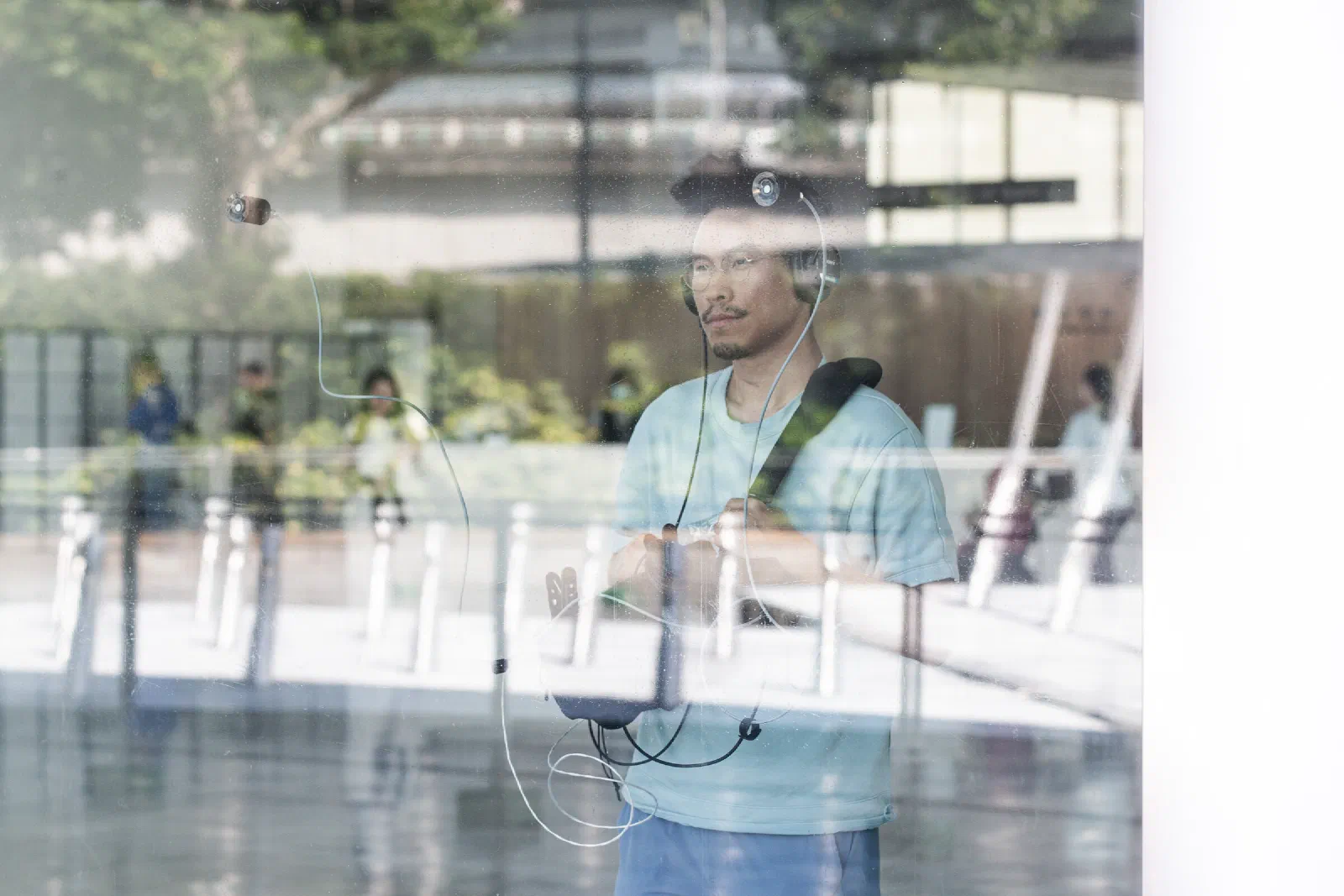A person wearing headphones stands behind a glass wall, with contact microphones attached to the glass by suction cups and trees and passers-by reflected in it.