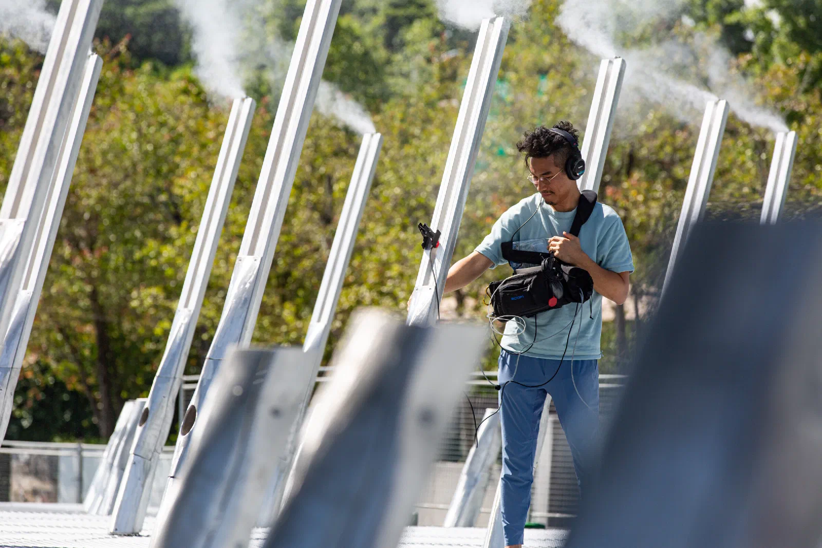 A person carrying a recorder bag walks between slanted white poles releasing plumes of mist, with green trees behind.