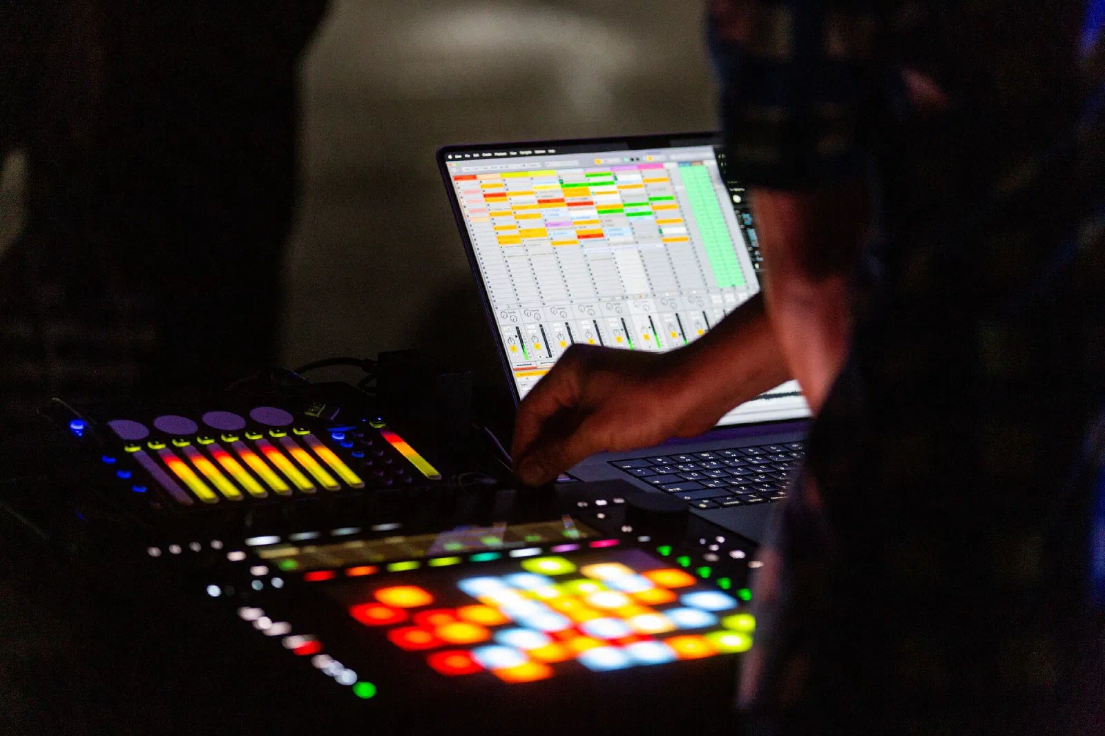 A hand adjusts a lit pad controller and fader unit in front of a laptop showing a clip-launching music software window, in a dark space.