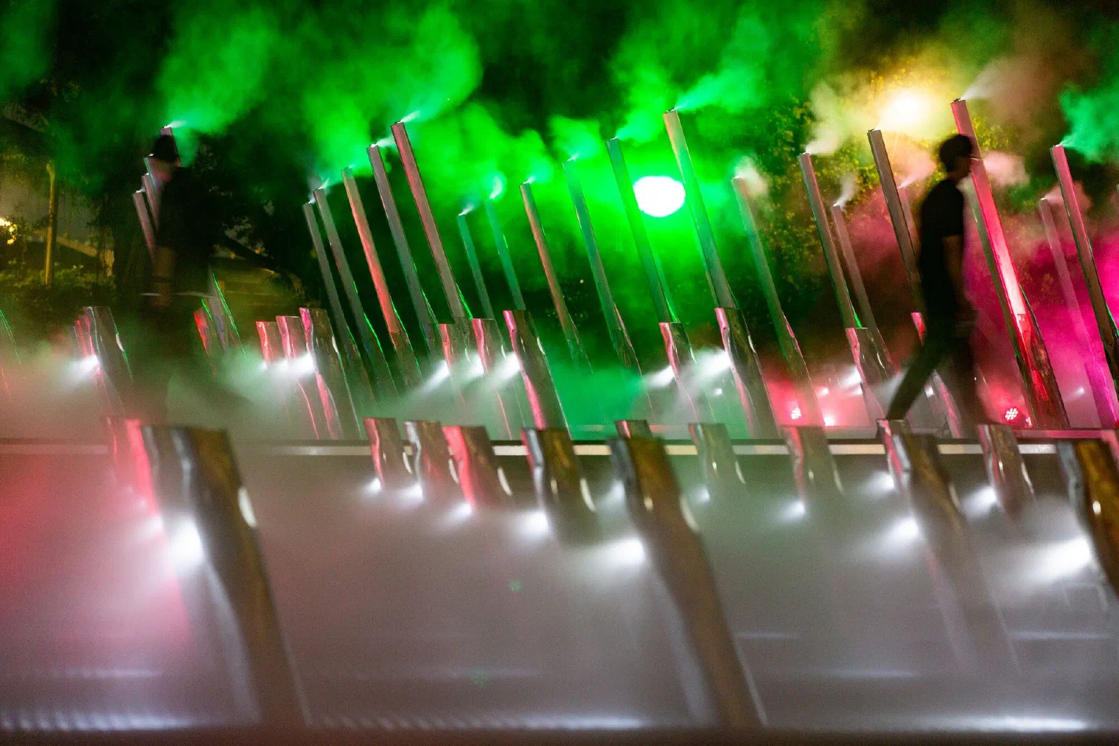 At night, slanted poles release green and pink mist while two blurred figures walk past in silhouette.